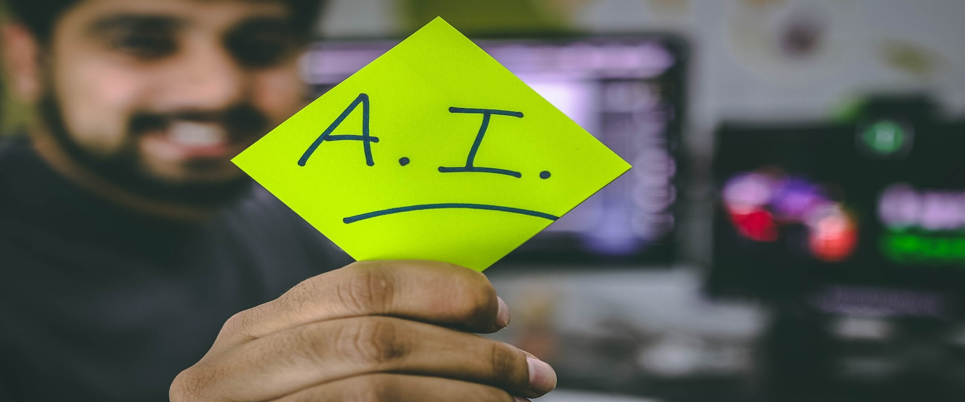 A smiling young man in the background is holding up a yellow post it note with the letters AI written on it.