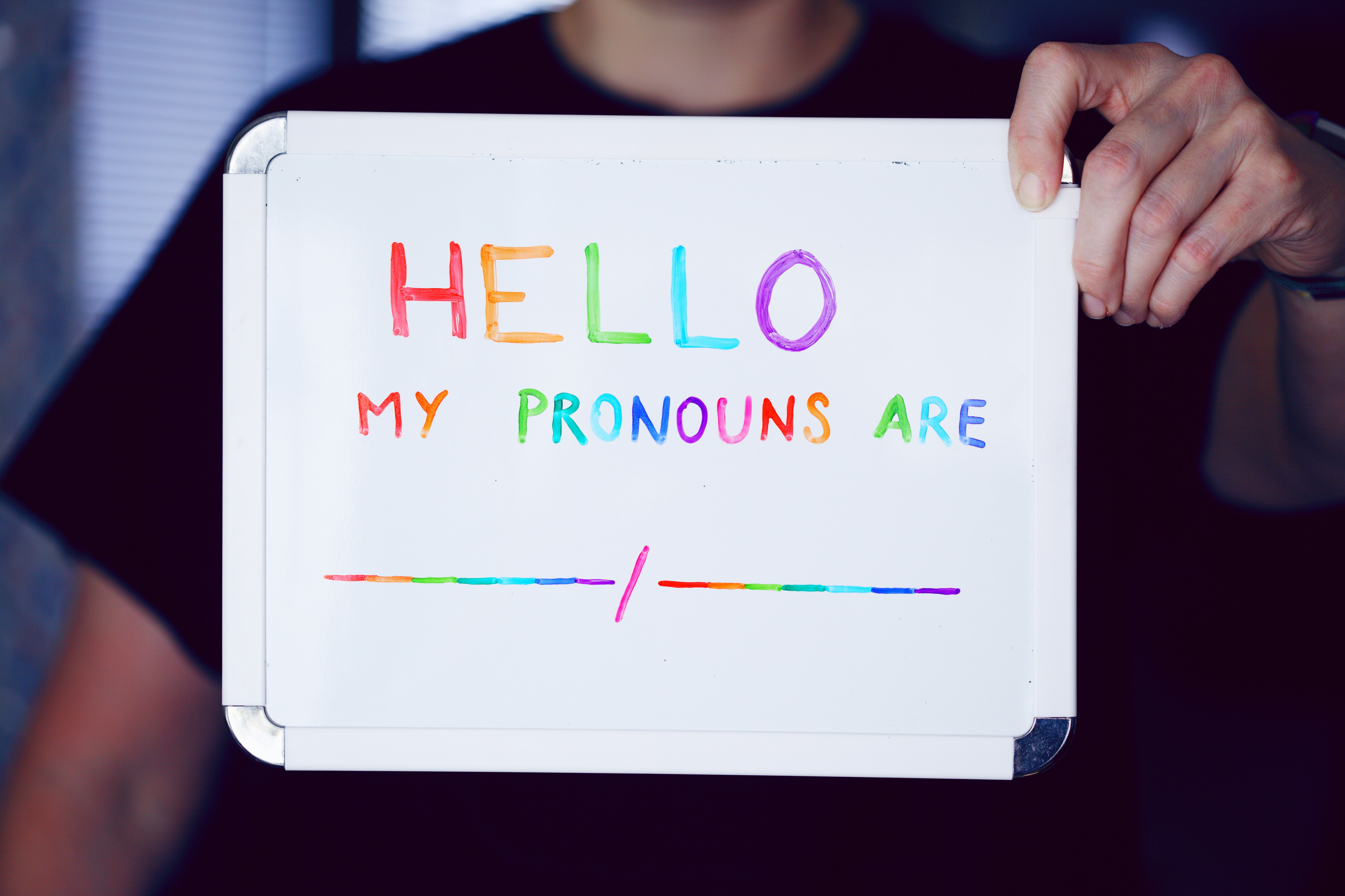 Person holding a whiteboard that says, in rainbow colors, Hello, My Prounouns Are" followed by blank lines