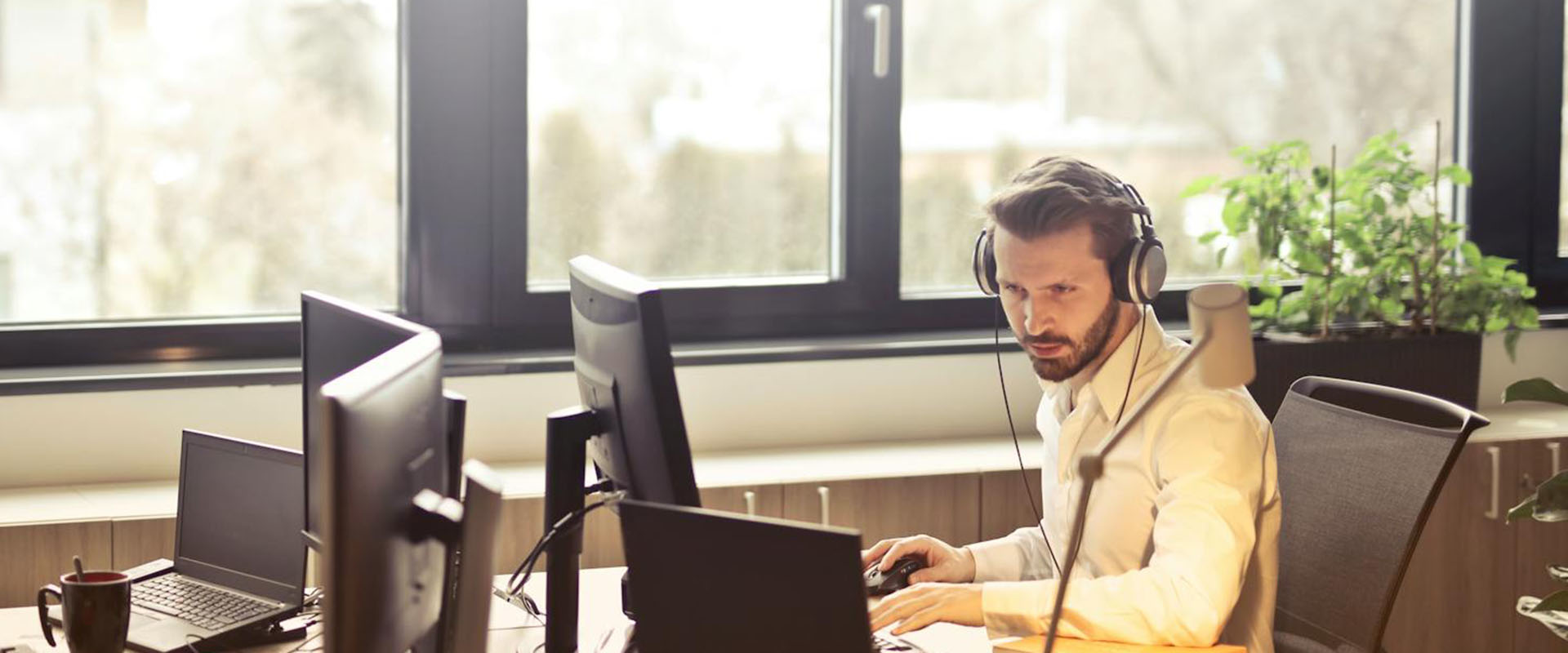 A man sitting at a desk working in front of two monitors. There are windows and trees in the background.